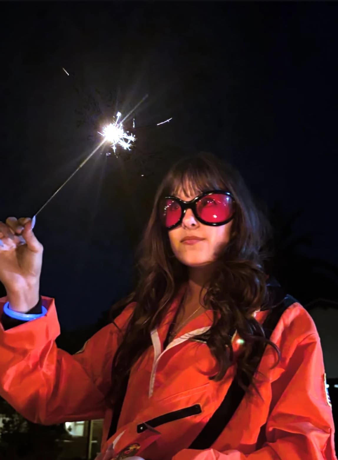 Synnove June at night holding a lit sparkler, wearing red sunglasses and an orange jacket
